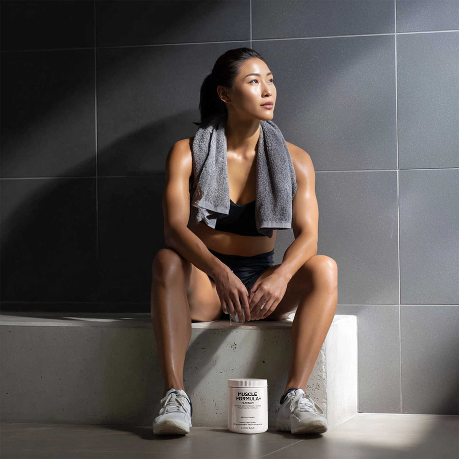 Woman sitting on a ledge with a towel draped over her shoulders, next to a container of Codeage Muscle Formula supplement