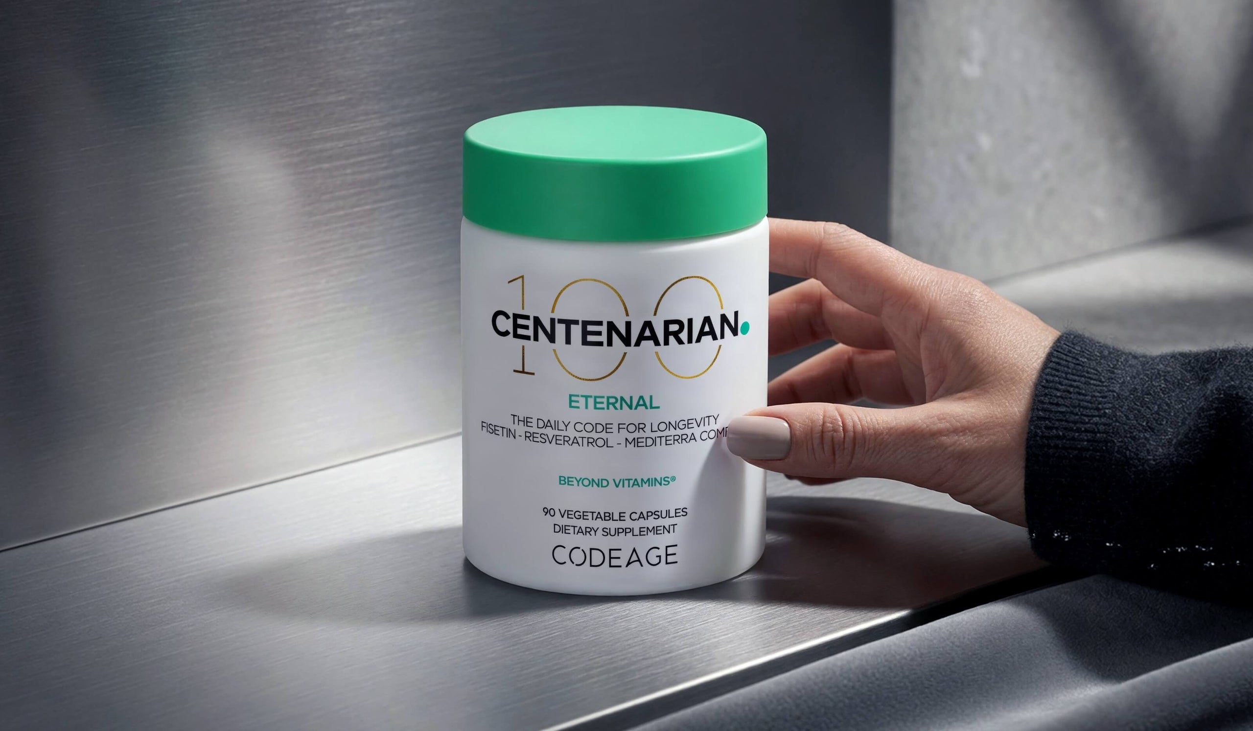 A hand holds a white Codeage Centenarian 100 Eternal dietary supplement bottle with a bright green cap on a brushed metal surface.